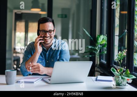 Portrait of young smiling cheerful entrepreneur in casual office making phone call while working with laptop Stock Photo