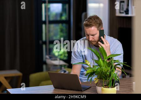 Portrait of casual pensive puzzled man using smart phone looking at laptop at home office having solving problems calling customer support Stock Photo