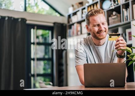 Happy cheerful smiling young adult man doing online shopping or e-shopping satisfied entrepreneur making online payment paying for service or goods ha Stock Photo