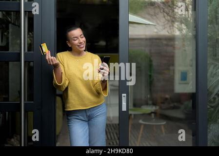 Portrait of casual person relaxed woman using phone online payment on home doorstep, female shopping online from home leaning door using credit card, Stock Photo