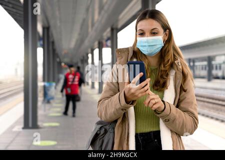 Beautiful young woman wearing medical mask ready to shows her sanitary passport on smartphone to get on the train at station outdoors Stock Photo