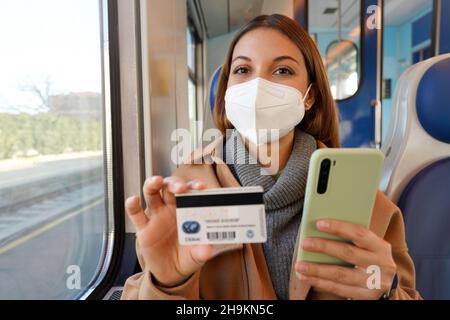 Happy confident woman with FFP2 KN95 protective mask traveling with train looking at camera holding credit card and smart phone Stock Photo