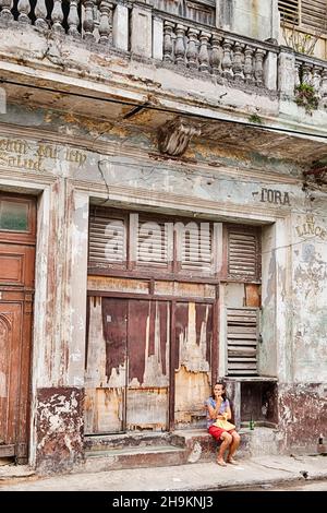 REGLA, CUBA - DECEMBER 22, 2019: An anonymous woman sits patiently in front of a shuttered old storefront in a scene typical of some of the quiet neig Stock Photo
