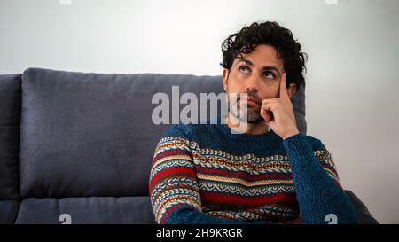 Portrait of handsome man wearing a winter sweater and thinking try solve problem pose on a couch at home interior. Worried caucasian man sitting on so Stock Photo