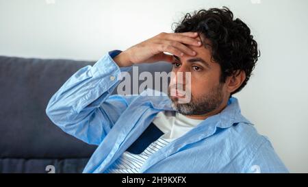 Portrait of handsome man wearing a shirt and feeling exhausted on a couch at home. Frustrated caucasian man holding head on hand and sitting on sofa o Stock Photo