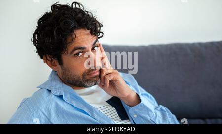 Portrait of cool and handsome man wearing a shirt and sitting on a couch at home interior. Good looking caucasian man on sofa of house. Handsome male Stock Photo