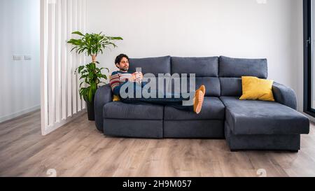 Portrait of caucasian man using smartphone lying down on couch at beautiful home interior. Caucasian man checks phone device on the sofa of house. Stock Photo