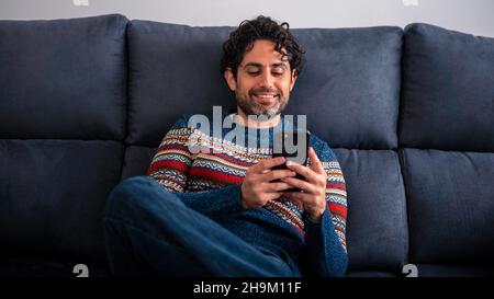 Happy handsome man smiling while using his smart phone and sitting on the couch at home. Confident bearded man relaxing and typing on smartphone at so Stock Photo