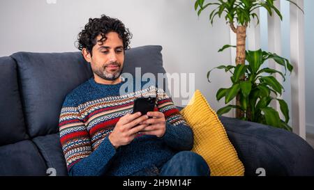 Cheerful handsome man using his smart phone while sitting on the couch at home. Confident bearded man is typing on smartphone at sofa of house. Stock Photo