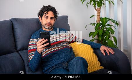 Portrait of caucasian man using smartphone sitting on couch at beautiful home interior. Caucasian man checks phone device on the sofa of house. Stock Photo