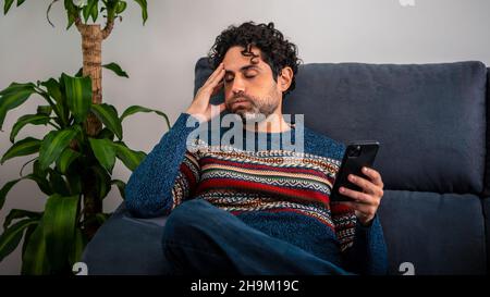 Male depression holding his forehead while having headache on couch home. Adult man using smart phone and thinking, having heavy thoughts, breake up w Stock Photo