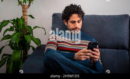 Portrait of caucasian man using smartphone sitting on couch at beautiful home interior. Caucasian man checks phone device on the sofa of house. Stock Photo