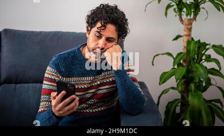 Male depression and sitting on couch home. Bearded adult man using his smart phone and thinking, having heavy thoughts, breake up with lover or financ Stock Photo
