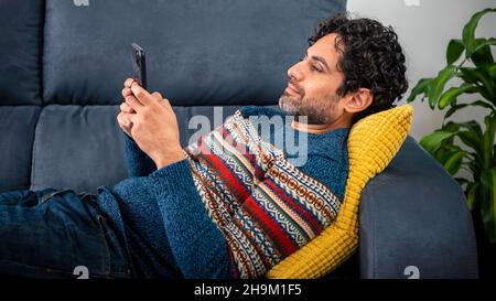 Portrait of caucasian man using smartphone lying down on couch at beautiful home interior. Caucasian man checks phone device on the sofa of house. Stock Photo