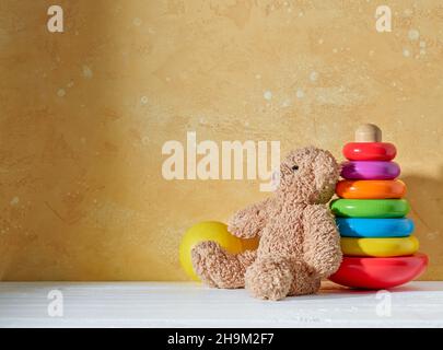 old teddy bear and colorful wooden toy pyramid on a white wooden shelf and yellow wall background Stock Photo