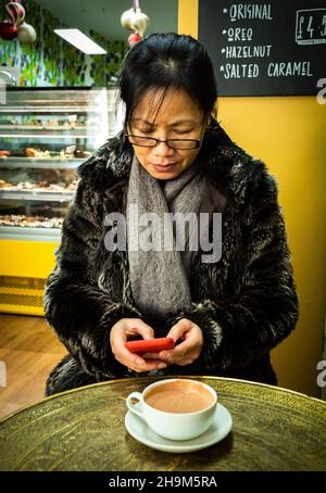 A Vietnamese woman uses her iphone in the artisan chocolate cafe called Cenu Cacao in King's Parade, Cambridge, UK. Stock Photo