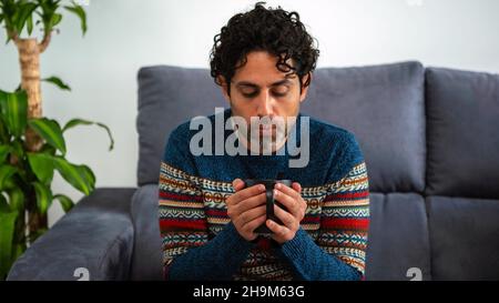 Portrait of cool and handsome man drinking morning expresso coffee and sitting on a couch at home interior. A person is resting and looking away. Stock Photo