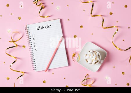 New year, Christmas or holiday wish list concept. Notepad, golden tinsel, and a cup of hot chocolate with marshmallows isolated on pink background Stock Photo