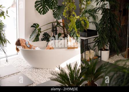 Relaxed young woman bathing in modern bathroom interior decorated with tropical plants Stock Photo