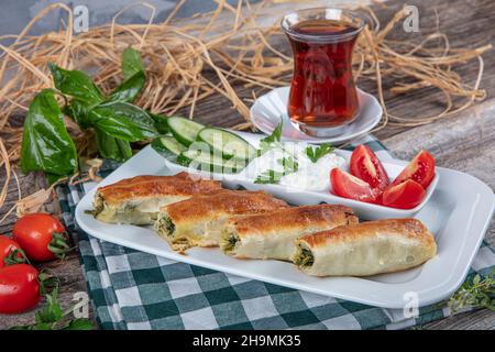 Turkish borek rolls with spinach on the white plate. A traditional Turkish pastry, rulo borek. Spinach Borek, tomato, cucumber and tea on the wooden t Stock Photo