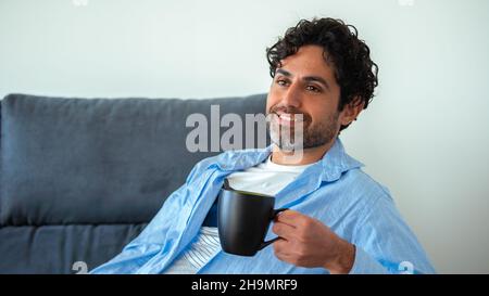 Portrait of happy man drinking morning expresso coffee while relaxing on couch and smiling. Cheerful caucasian guy having cafe and feeling confident i Stock Photo