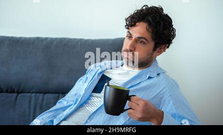 Handsome male model feels tired after work and having a cafe. Mature man drinking morning expresso coffee while having headache while sitting on couch Stock Photo