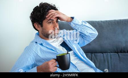 Portrait of handsome man drinking morning expresso coffee and feeling exhausted on a couch at home. Frustrated caucasian man having cafe while holding Stock Photo