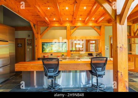 Island with stainless steel countertop and oiled pine wood  bar with black plastic and leather high back chairs in kitchen inside timber frame home Stock Photo