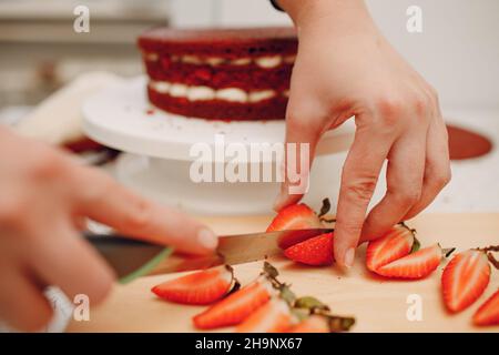 Pastry chef makes delicious red velvet cake. Cutting strawberry and decorating dessert. Stock Photo
