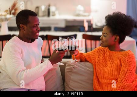Happy african american couple drink red wine on sofa, celebrating anniversary in quarantine at home Stock Photo