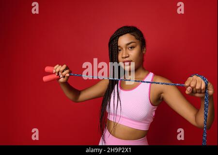 African American pretty woman in pink sportswear holds a jumping rope in front of her and showing it to a camera confidently looking at it, posing ove Stock Photo