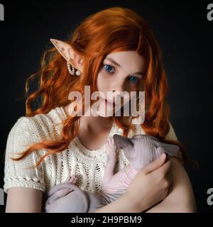 Close-up view of ginger young woman cosplayer elf in white knitted dress, with long wavy hair, blue eyes, cradling Sphinx kitten and looking away Stock Photo
