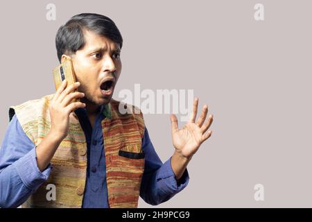 A young Indian male in blue dress talking over phone with agitated expression on gray background Stock Photo