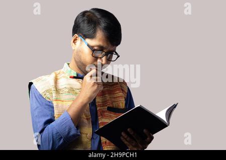 A young Indian male in ethnic dress thinking with pen and notebook on gray background Stock Photo