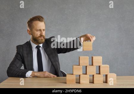 Business leader sitting at office table and building pyramid out of wooden cubes Stock Photo