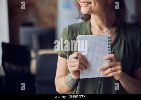 Cropped shot of young woman in casual wear holding empty notebook and pen in her hands and smiling aside. Interior designer or architect thinking Stock Photo