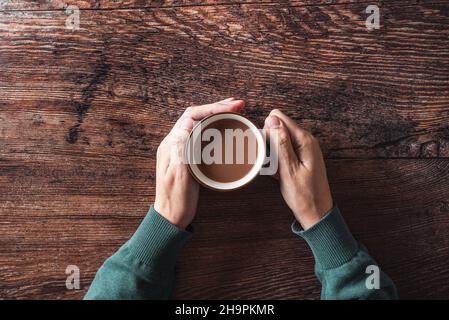 From above of crop unrecognizable male touching warm mug with hot coffee with milk while sitting at wooden table Stock Photo