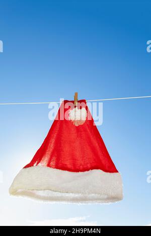 A Santa Claus hat hung on a clothesline and fastened with a clothespin. In the background is the blue sky with some small clouds. Stock Photo