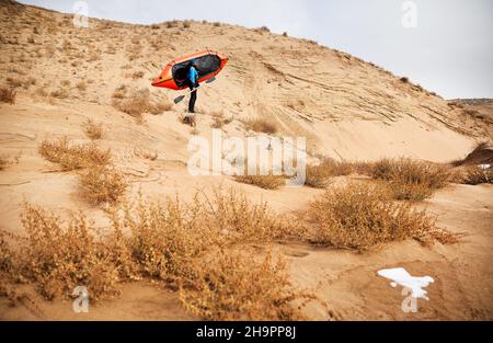Old man with black beard in blue jacket holding orange raft sport boat at sand dune in the desert background. Adventure and travel concept. Stock Photo