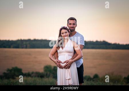 Young couple expecting new life. Pregnant woman with her husband holding belly. Sky background after sunset Stock Photo