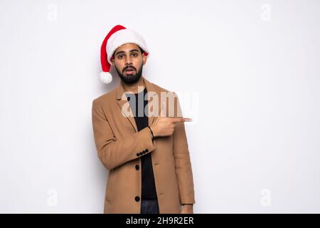 Young man in Santa Claus hat is surprised and points fingers towards isolated on white background Stock Photo