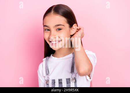 Photo portrait of small schoolgirl smiling touching hair in overall isolated on pastel pink color background Stock Photo