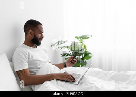 Serious millennial african american bearded guy sit on bed in bedroom interior, working on laptop Stock Photo