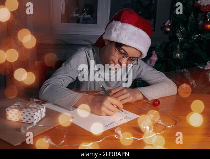 A boy wearing glasses, red cap like Santa writes letter to Santa on wooden table near Christmas tree. Selective blurred focus. Bright light bokeh. Chr Stock Photo