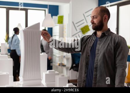 Inspired architect holding pen measuring skyscraper foam model standing next to desk with residential maquette. Engineer looking for inspiration in modern architectural office. Stock Photo