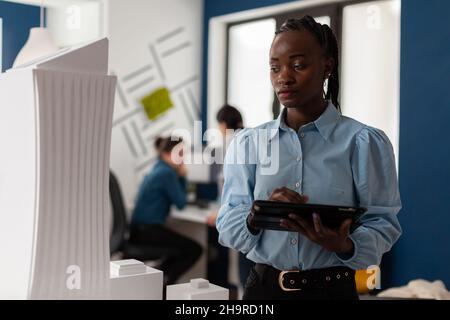 African america architect looking at scale foam model of modern building while holding tablet with blueprints. Architectural engineer focused on the design of 3d skyscraper maquette. Stock Photo