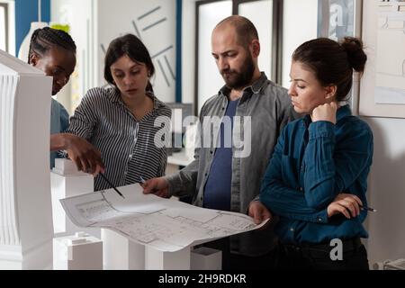 Team of architects analyzing blueprints for building project standing in front of table with real estate maquette. Professional engineers reading construction plans for urban development. Stock Photo