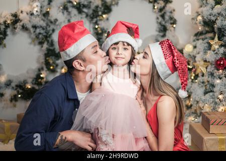 Christmas studio family shoot in red Christmas hats. New Year photo of happy mixed family with cute daughter. Mother and father kissing their daughter Stock Photo