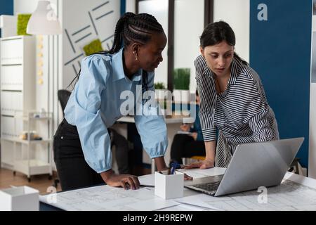 Team of two professional women architects working on laptop computer with construction plans on desk in architectural office. Engineers designing blueprints for real estate project. Stock Photo