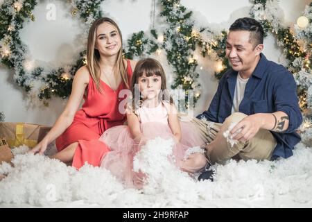 Christmas studio family shoot. New Year's photo of happy family. Christmas wreaths and artificial snow. Mother, father and cute daughter in pink dress Stock Photo